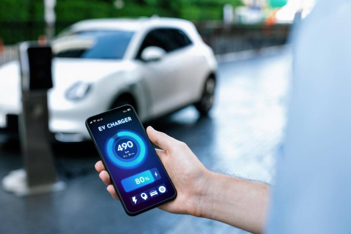 A hand holds a smartphone displaying EV charger details: 490 km range and 80% charge, with a white electric car parked nearby.