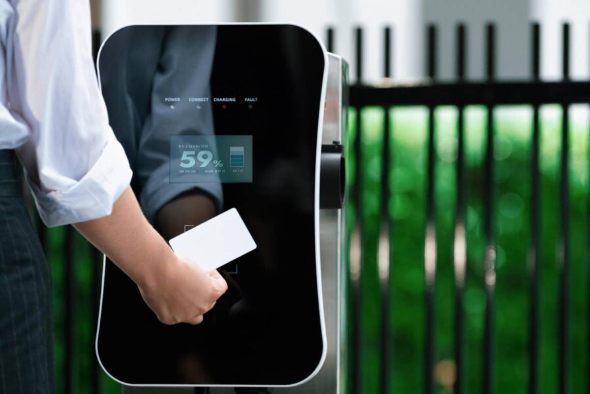 A person holds a card near a sleek EV charging station displaying battery status and connection options, against a green backdrop.