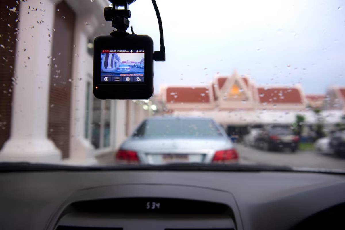 Dashboard camera recording through a rain-speckled windshield, capturing a parking lot scene with nearby vehicles and buildings.