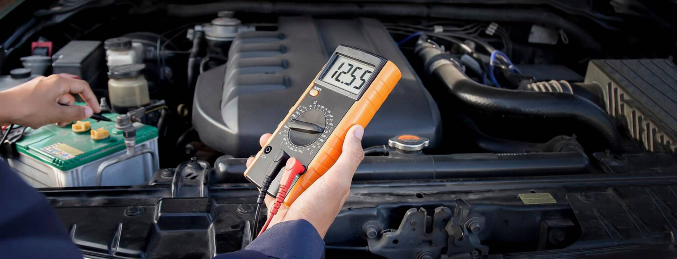 A mechanic holds a multimeter, testing a car battery, displaying a reading of 12.55 volts, with an engine in the background.