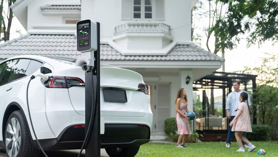 A white electric car is charging at home, with a charging station displaying 60% battery, while a family plays nearby in a garden.