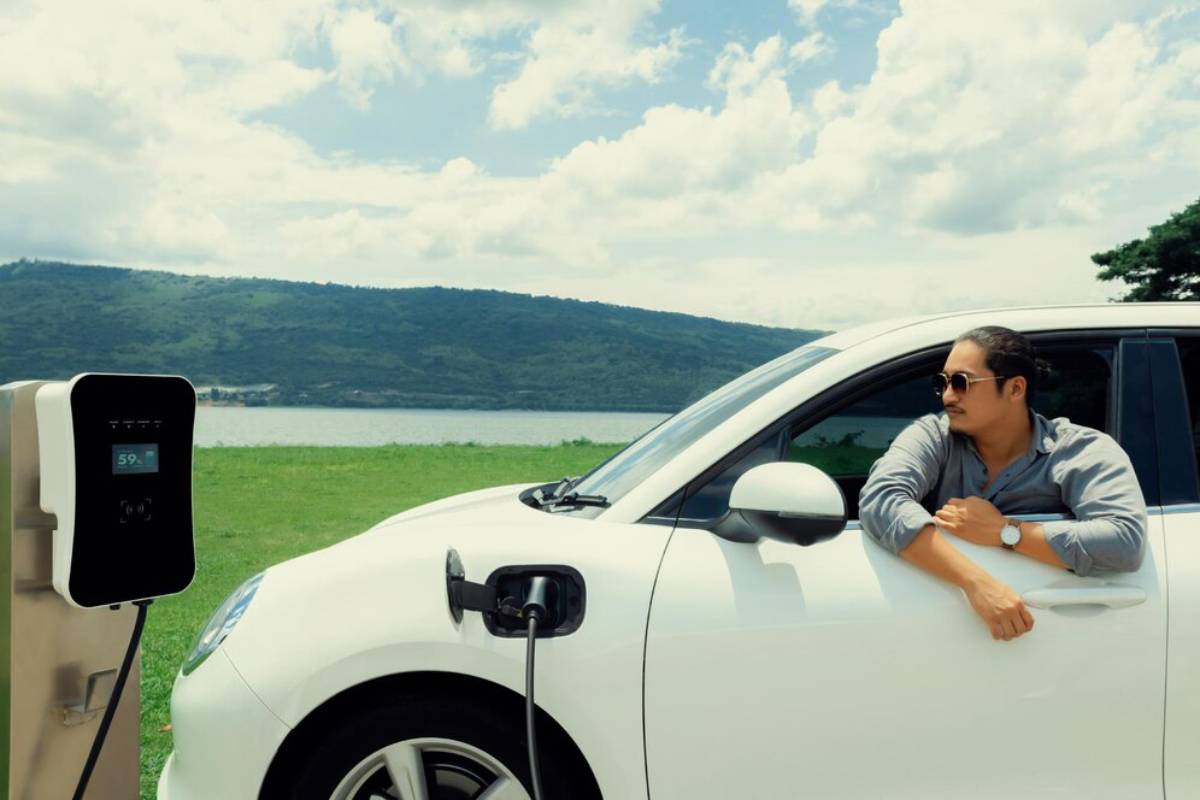 A white electric car is plugged into a charging station, with lush green hills and a lake visible in the background under a cloudy sky.
