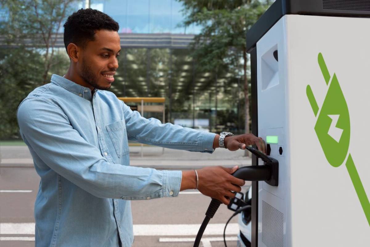 A person connects an electric vehicle charger to a charging station, with modern buildings and greenery in the background.
