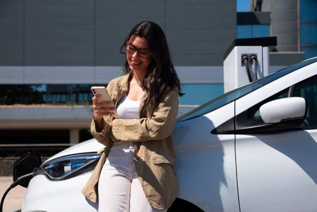 A woman in a beige jacket leans against a white electric car, checking her phone while charging at a modern station.