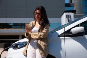 A woman in a beige jacket leans against a white electric car, checking her phone while charging at a modern station.