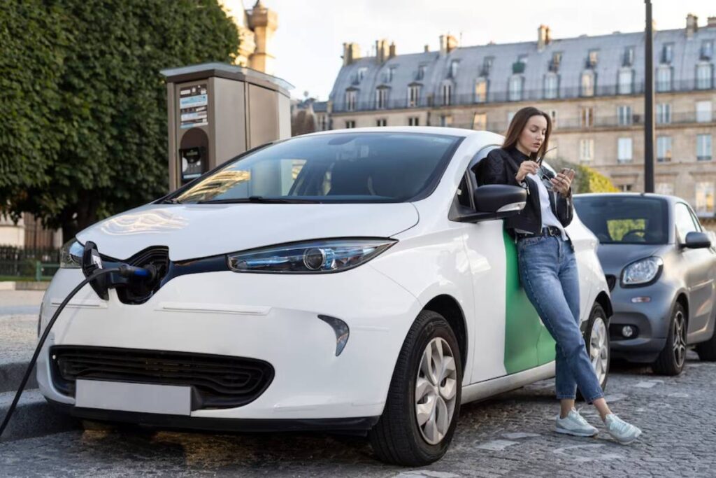 A white electric car is being charged at a station, while a person leans against it, looking at their phone, in an urban setting.