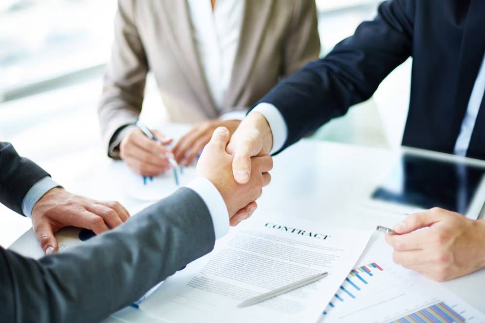 Two business people shaking hands over a contract, with a woman taking notes in the background.