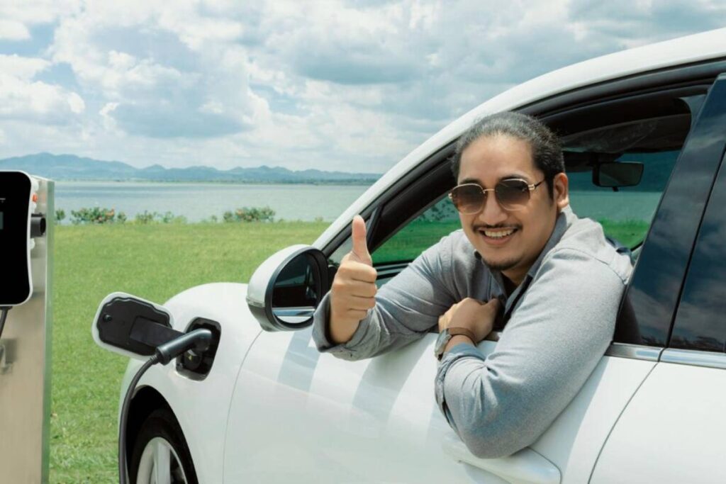 A person gives a thumbs up from the driver's side of a white electric vehicle, parked near a scenic lake under a cloudy sky.