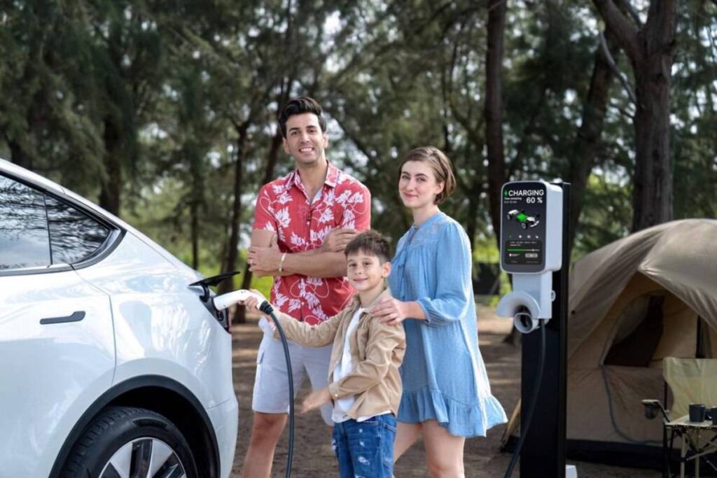 A family beside a white electric vehicle charging at a station in a forested camping area, with a tent in the background.