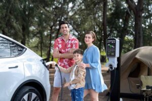 A family beside a white electric vehicle charging at a station in a forested camping area, with a tent in the background.