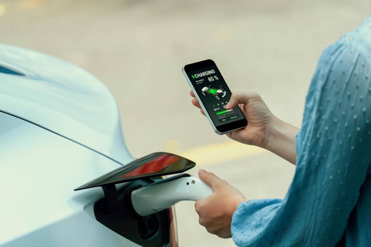 A person checks their smartphone displaying a 60% charge status while connecting an electric vehicle to a charging station.