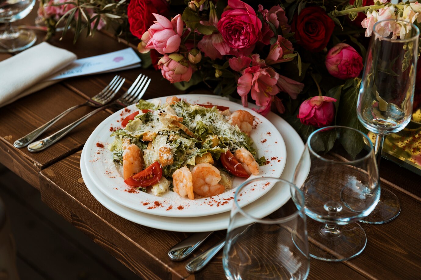 A gourmet shrimp salad served on a white plate beside a floral centerpiece and wine glasses on a wooden table.