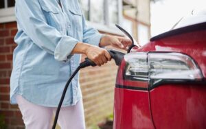 A woman plugs a charging cable into the rear of a red electric car, standing in front of a brick home.