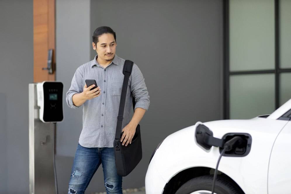 A man stands near an electric vehicle, checking his phone while the car is plugged into a charging station.