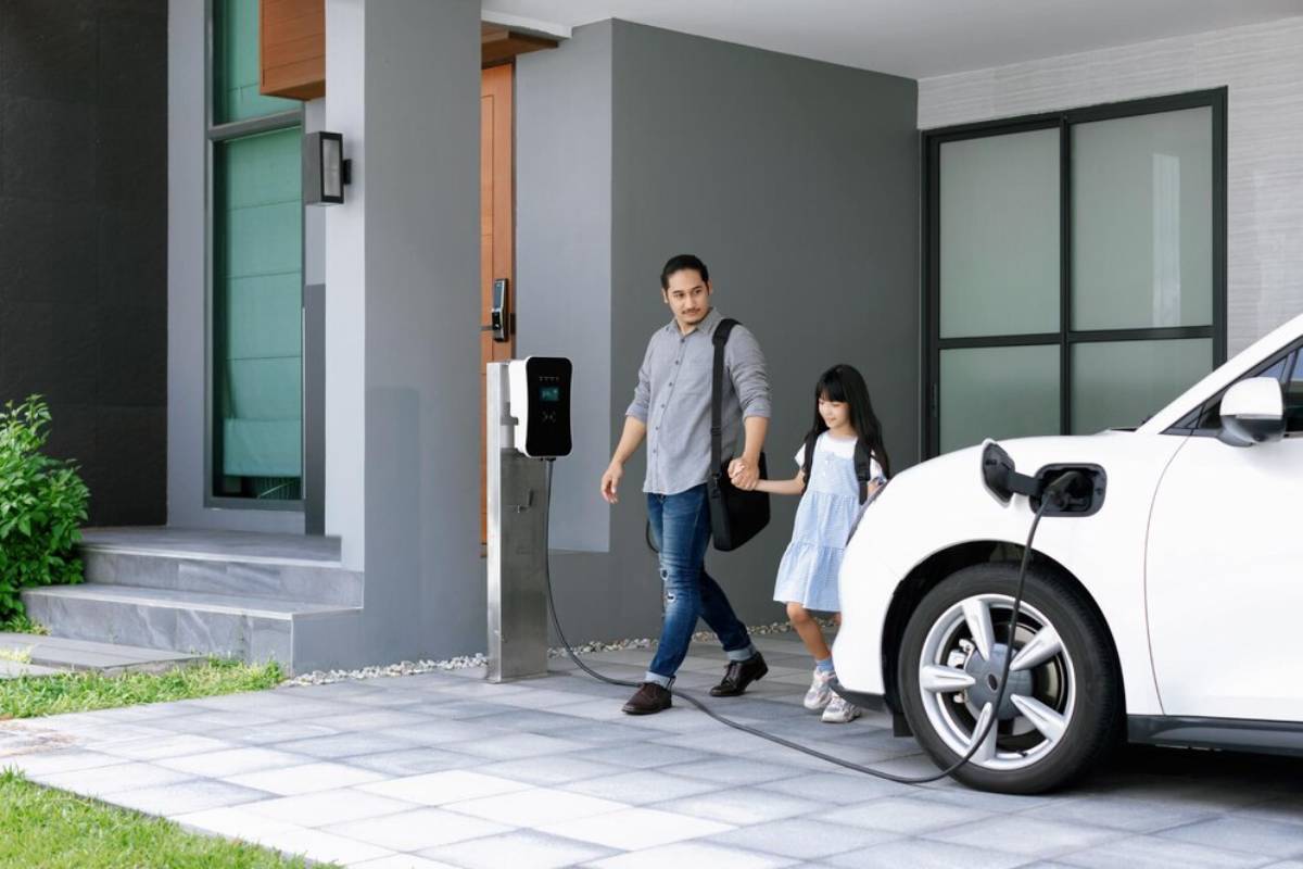 A man and a girl walk hand-in-hand towards a white electric car charging at a home, with green grass and modern architecture in view.
