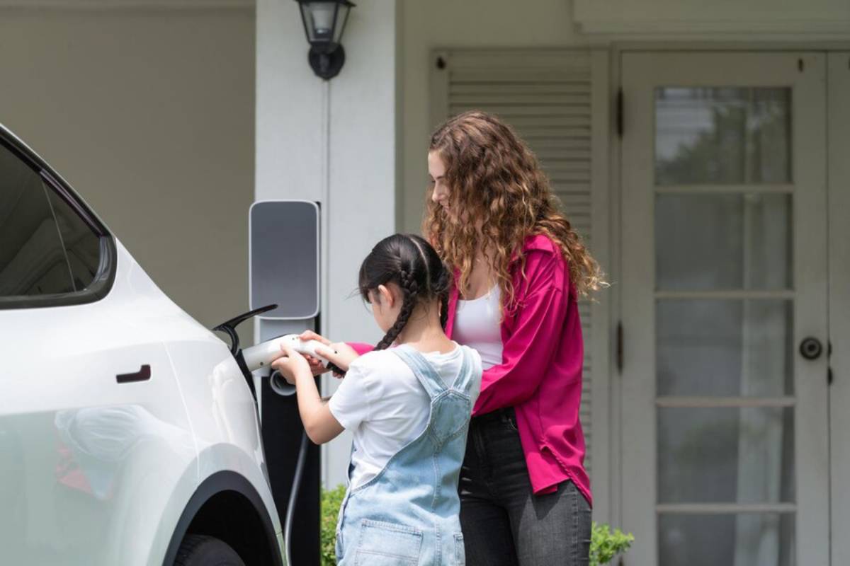 A woman and a child are connecting a charging cable to a white electric vehicle outside a modern home.