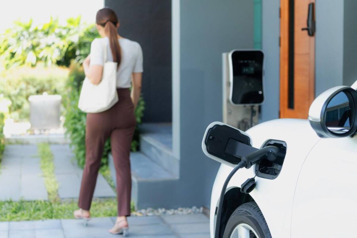 A woman with a tote bag walks away from a white electric vehicle charging at home, surrounded by greenery and modern architecture.