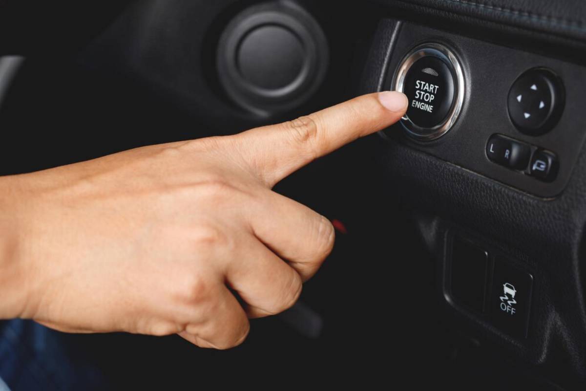 A hand pressing the start/stop button in a car, ready to ignite the engine. Control buttons are visible on the dashboard.