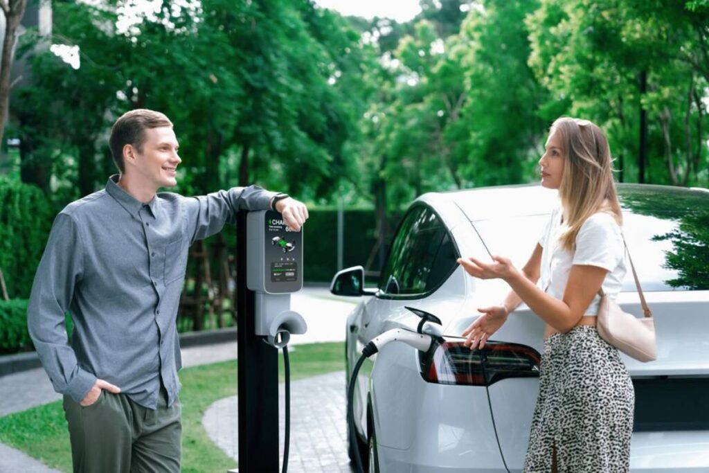 A man and woman converse near an electric vehicle charging station, with a sleek car plugged in under lush greenery.