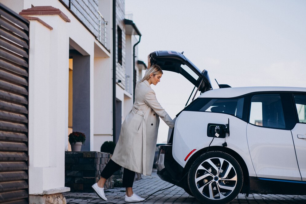 A person in a long cream coat is loading items into the trunk of a white electric car parked outside a modern house.