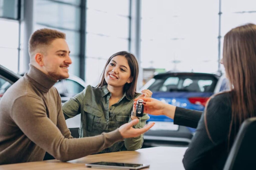 A car dealership scene where a woman hands over car keys to a man, while another woman gestures towards him, showcasing a transaction.
