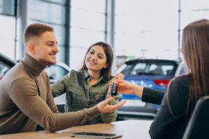 A car dealership scene where a woman hands over car keys to a man, while another woman gestures towards him, showcasing a transaction.