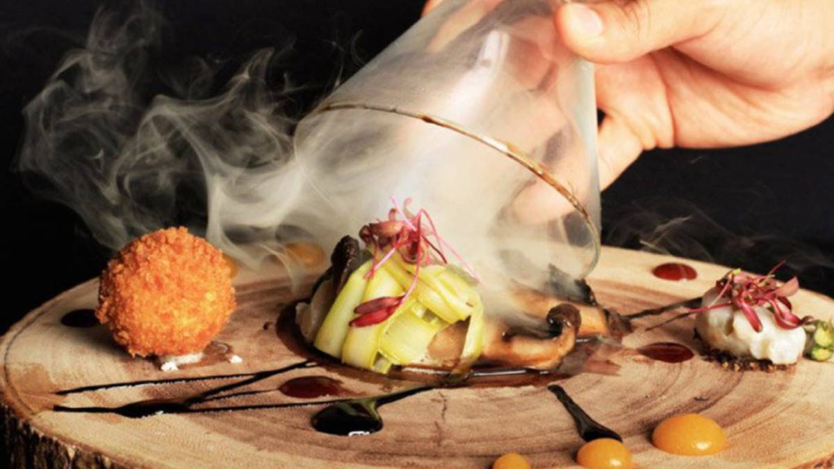 A hand lifting a smoke-filled glass dome from a creatively presented dish on a wooden platter.