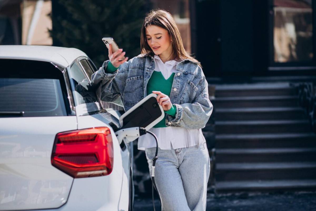 A woman in a denim jacket uses her phone while charging a white electric car. Steps and greenery are visible in the background.