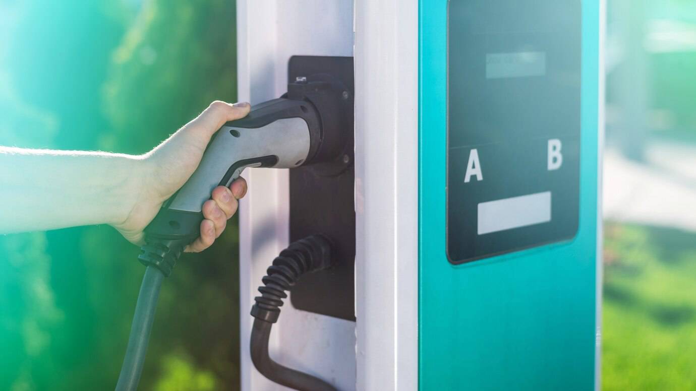 A hand connects an electric vehicle charging plug to a charging station, with a bright background and two labeled charging ports visible.
