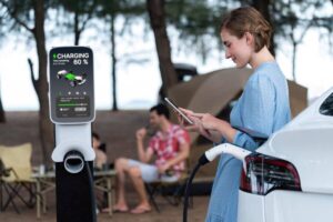 A woman in a blue dress checks her phone while her electric vehicle charges at a station displaying 60% battery.