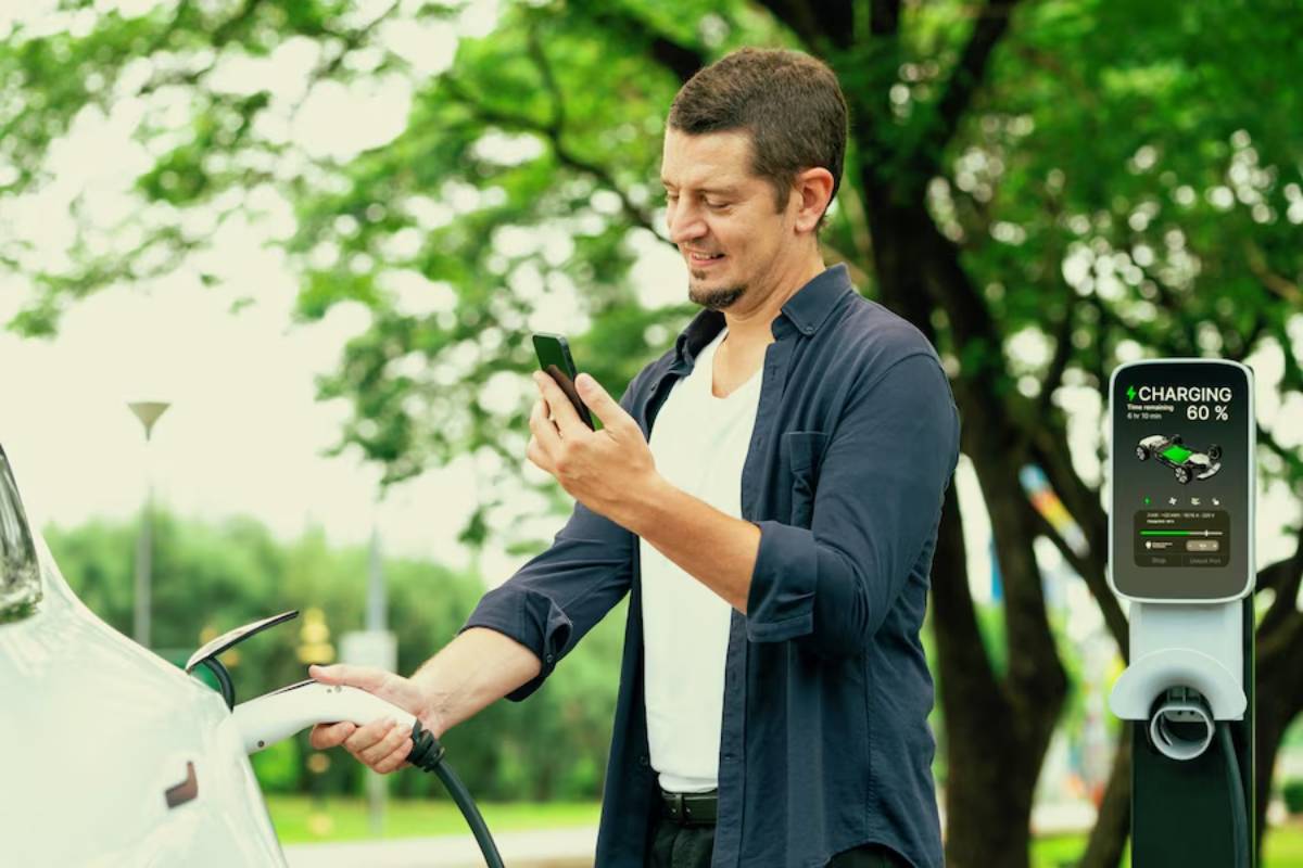 A person stands beside a charging electric vehicle, holding a smartphone, with a charging station displaying 60% charged. 