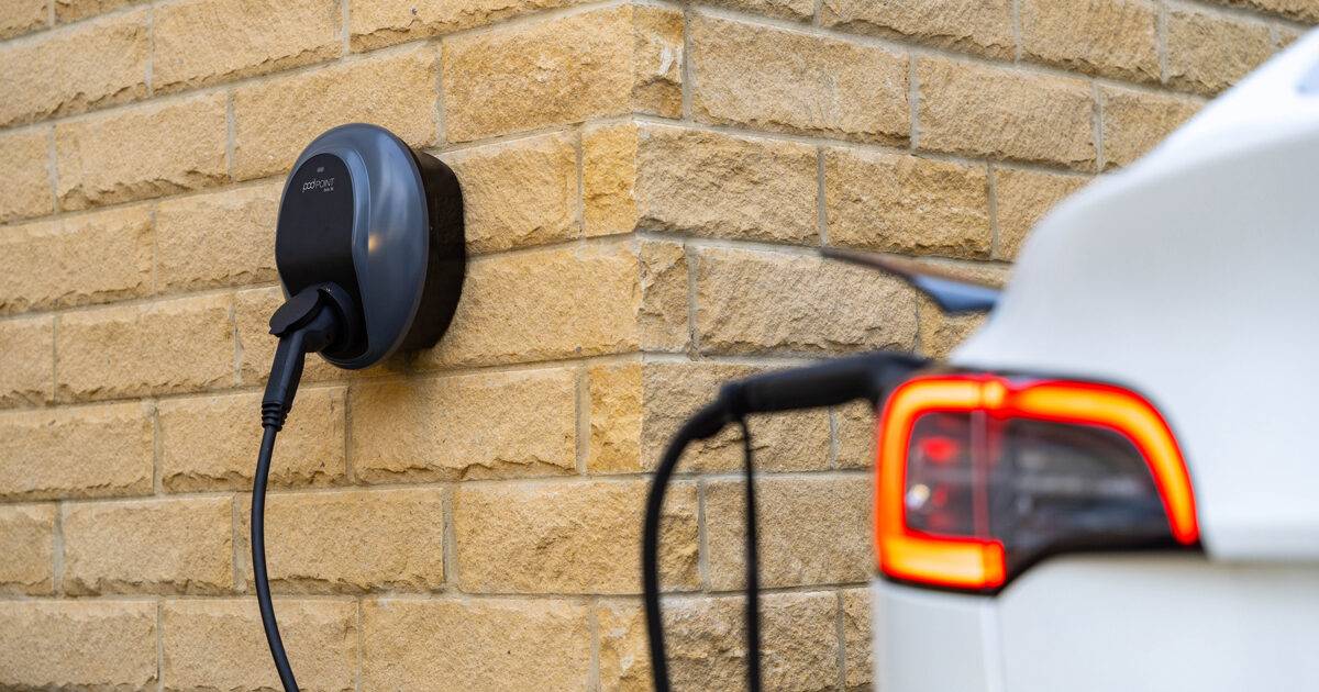 A white electric car is plugged into a wall-mounted charging station, set against a textured stone wall.
