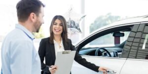 A car saleswoman in a black suit holds a clipboard while discussing details with a customer near a shiny white car.