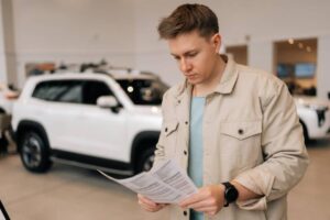 A person in casual attire examines paperwork while looking at a white SUV in a stylish car dealership.