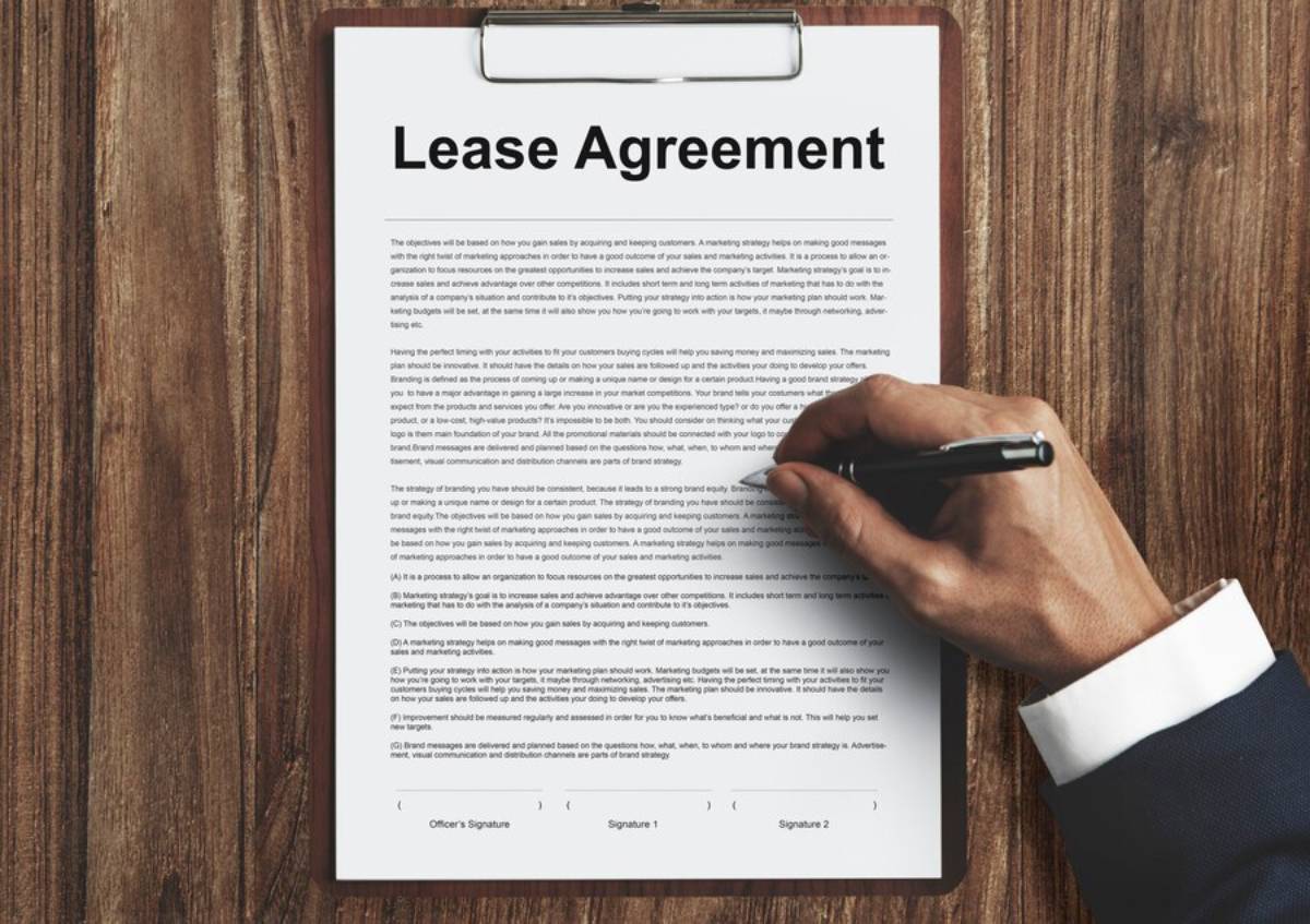 A hand signing a lease agreement document on a clipboard, with a focus on the bold header. The background is a wooden surface.