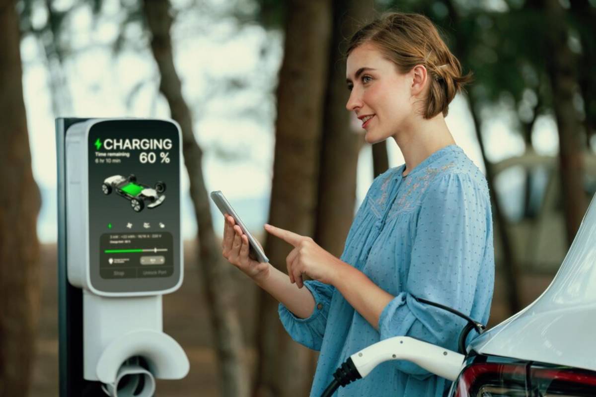 A woman in a blue blouse uses her smartphone while an electric vehicle charges at a station, displaying 60% battery status.