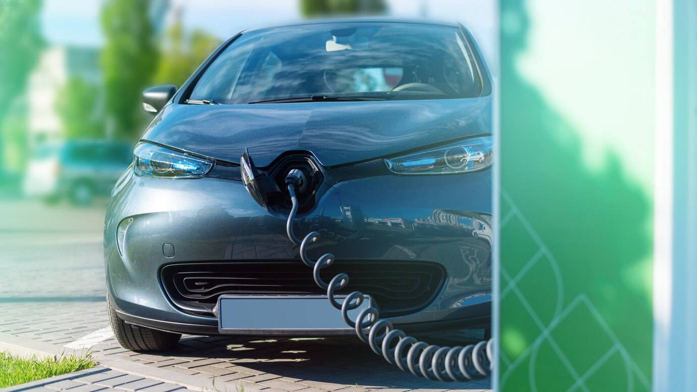 A gray electric car charging at a station, with a coiled charging cable connected to its front.