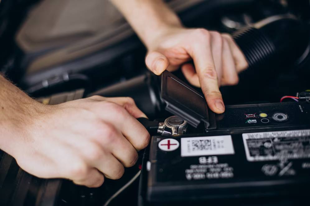 A person working on a car battery, opening a cover and adjusting connections, with an open hood in the background.