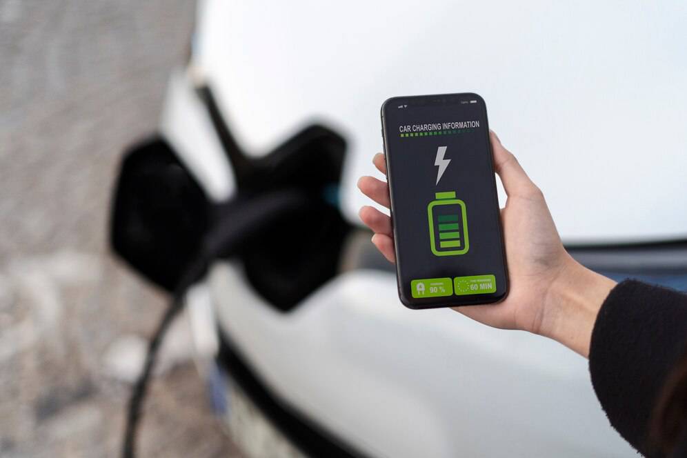 A hand holding a smartphone displays car charging details, showing battery percentage and charging time, next to an electric vehicle.