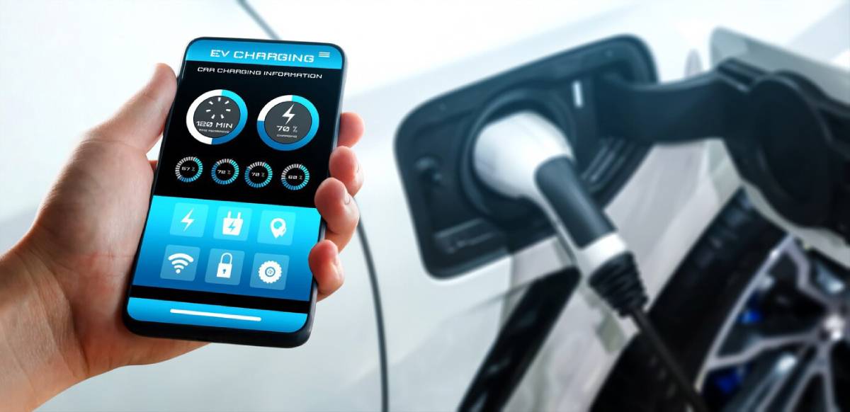 A hand holds a smartphone displaying EV charging status, while the phone is connected to an electric vehicle's charging port.