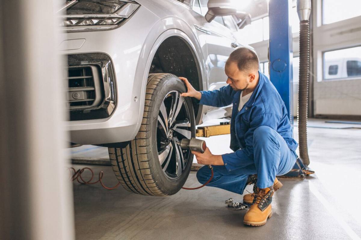 A mechanic in blue coveralls uses a power tool to remove a tire from a lifted silver SUV in a bright garage.