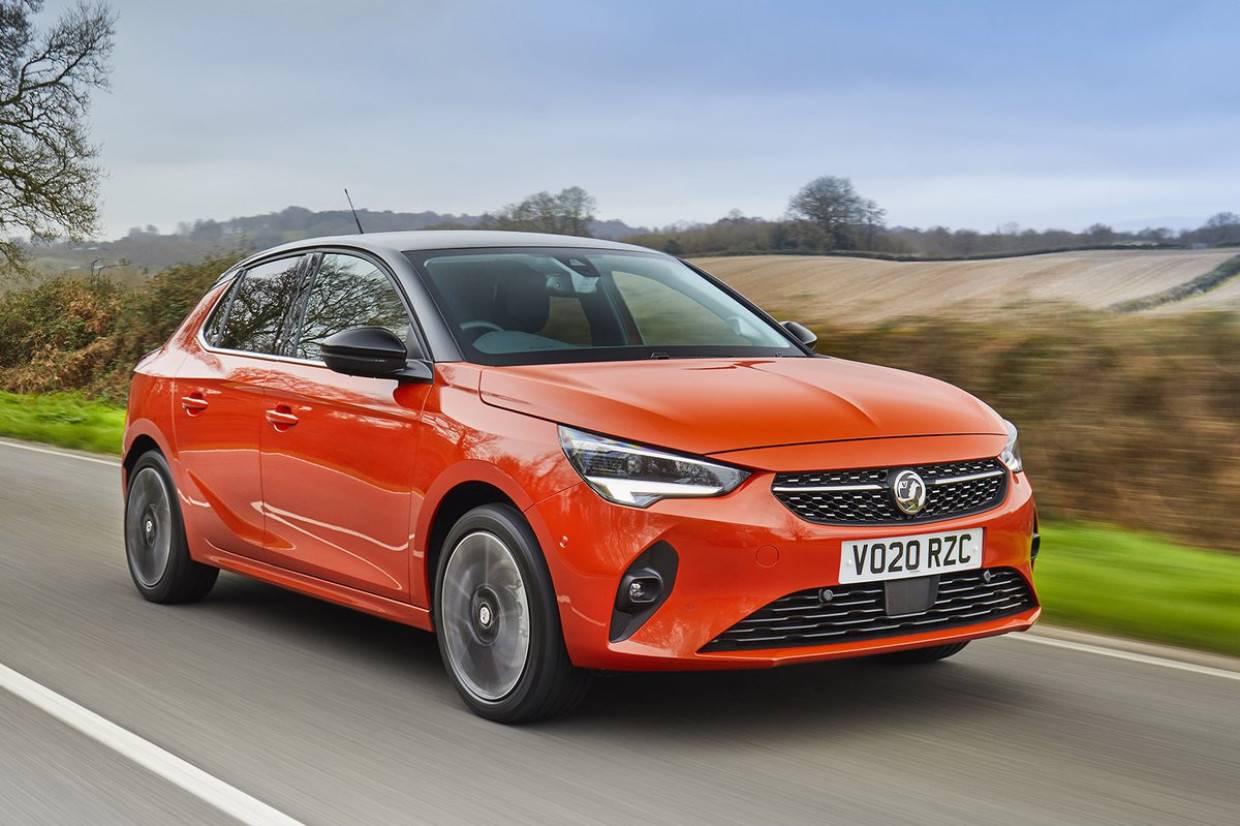 An orange Vauxhall Corsa drives on a rural road, showcasing sleek design against a backdrop of fields and trees.