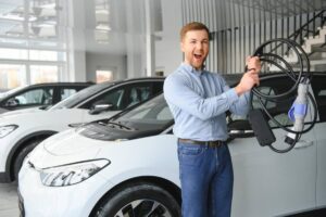 A man stands in an electric vehicle showroom, holding a charging cable alongside white electric cars in the background.