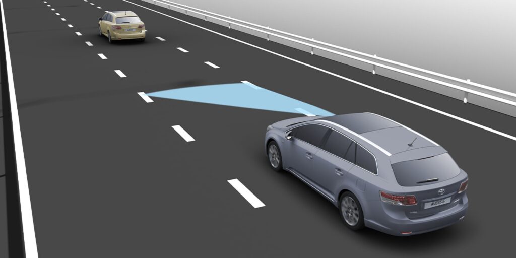 A gray Toyota Avensis on a highway, detecting a nearby yellow car with a blue lane departure warning illuminated.