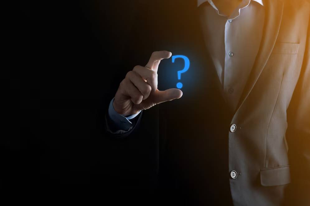 A suited man holds a glowing blue question mark between his fingers against a dark background, symbolizing inquiry or curiosity.