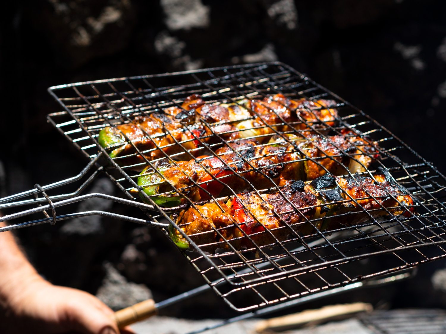 Grilled skewers with vegetables and meat on a barbecue rack held by a person's hand outdoors.