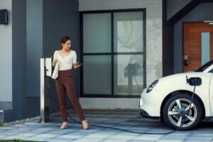 A woman in a white top and brown pants stands by an electric vehicle charging station outside a modern home, holding a phone.