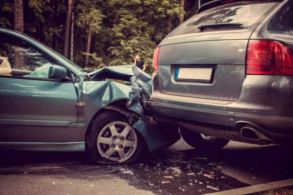 A damaged light blue car has crashed into the back of a silver SUV, with debris scattered on the road and trees in the background.