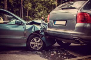 A damaged light blue car has crashed into the back of a silver SUV, with debris scattered on the road and trees in the background.