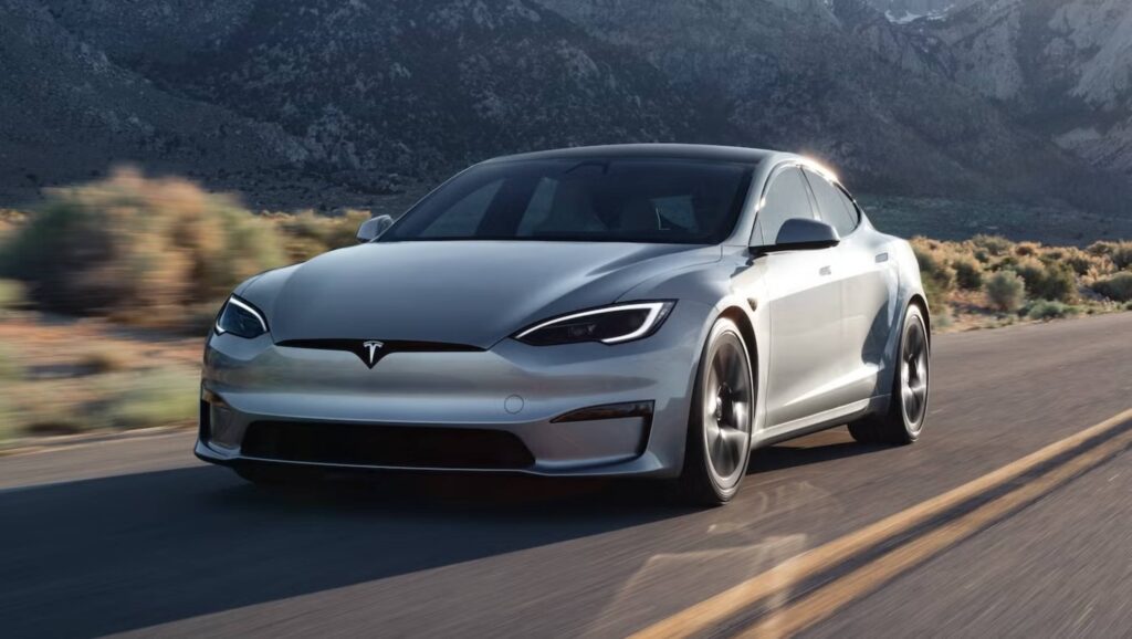 A sleek silver Tesla Model S drives on a winding road, with mountains and vegetation in the background under a clear sky.